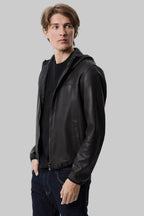 front side pose Full-length studio shot of a male model wearing a modern, slim-fit black leather hooded bomber jacket over a black t-shirt and dark blue jeans against a solid white background.