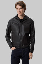 front pose Full-length studio shot of a male model wearing a modern, slim-fit black leather hooded bomber jacket over a black t-shirt and dark blue jeans against a solid white background.