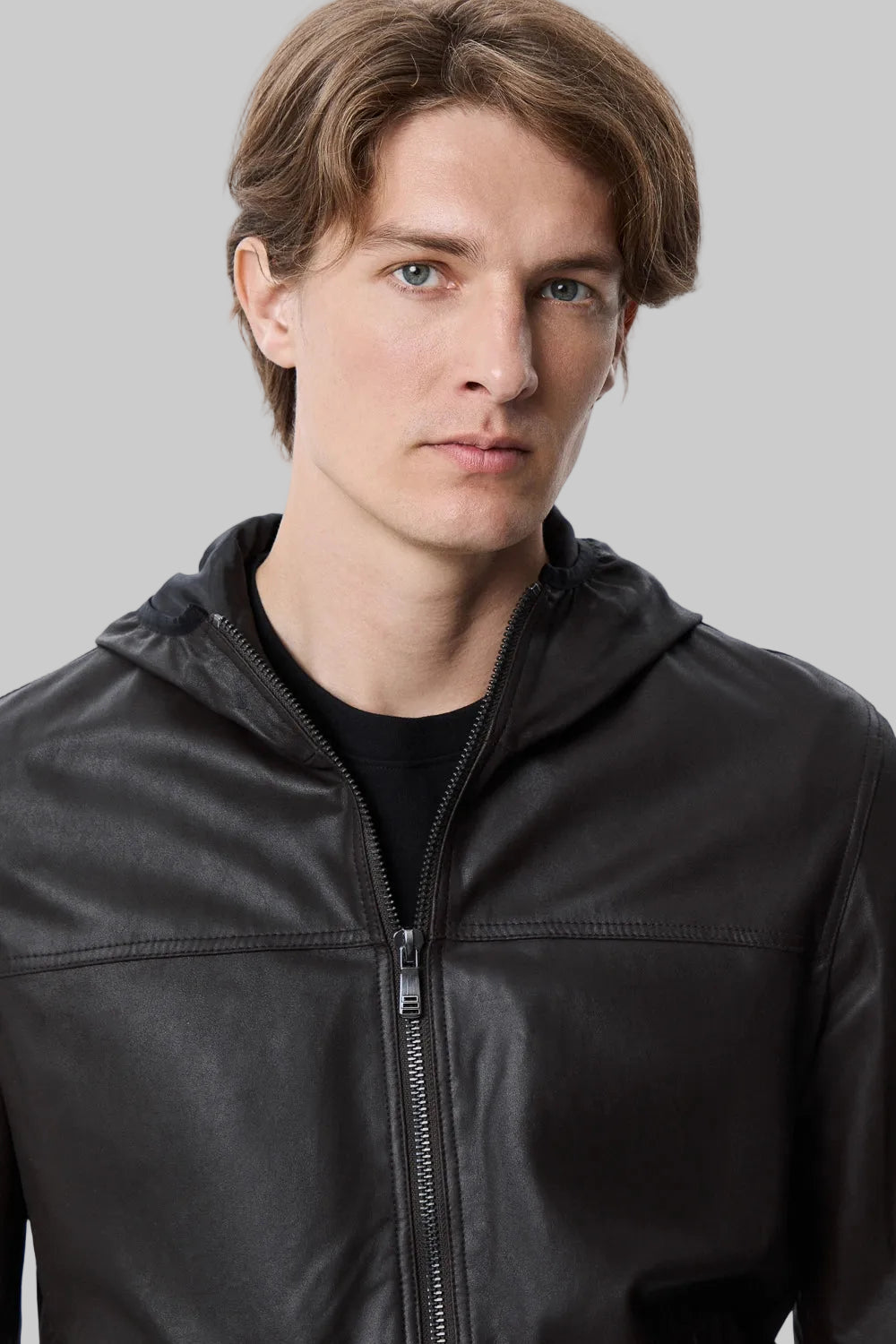 near posse Full-length studio shot of a male model wearing a modern, slim-fit black leather hooded bomber jacket over a black t-shirt and dark blue jeans against a solid white background.