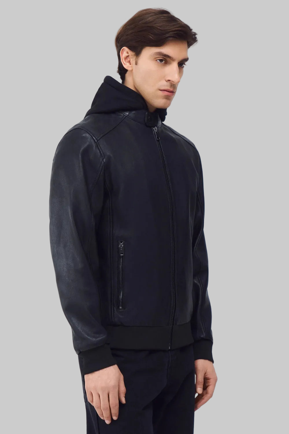 Front studio shot of a man wearing a slim-fit black leather bomber jacket with a snap-tab collar and zippered pockets, styled with dark pants against a white background.