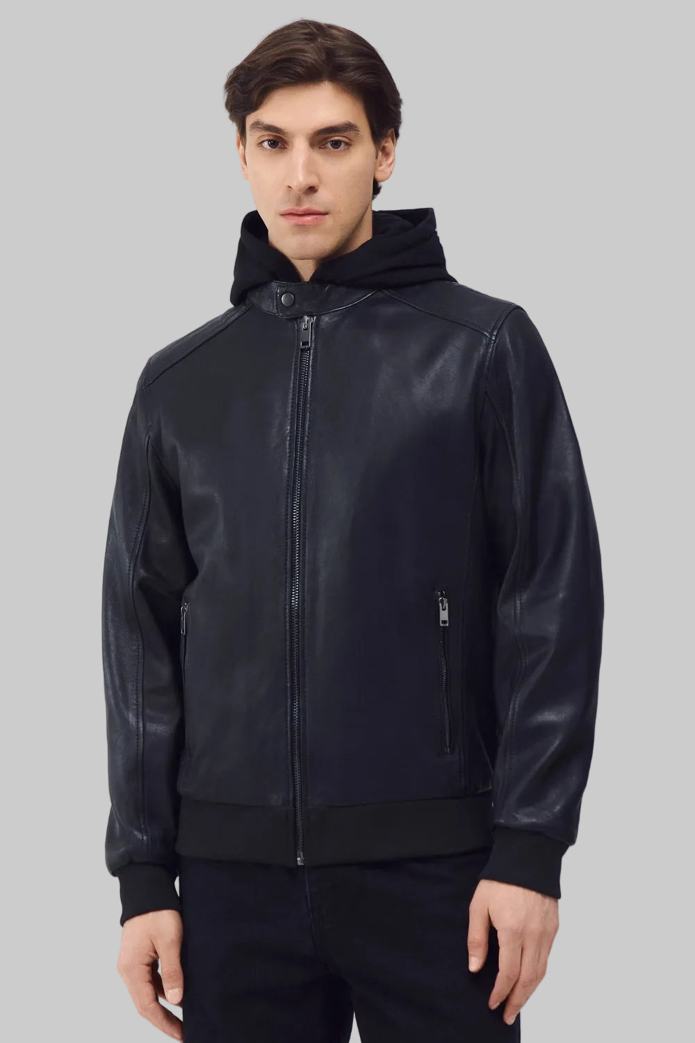 Front studio shot of a man wearing a slim-fit black leather bomber jacket with a snap-tab collar and zippered pockets, styled with dark pants against a white background.
