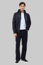 Front studio shot of a man wearing a slim-fit black leather bomber jacket with a snap-tab collar and zippered pockets, styled with dark pants against a white background.