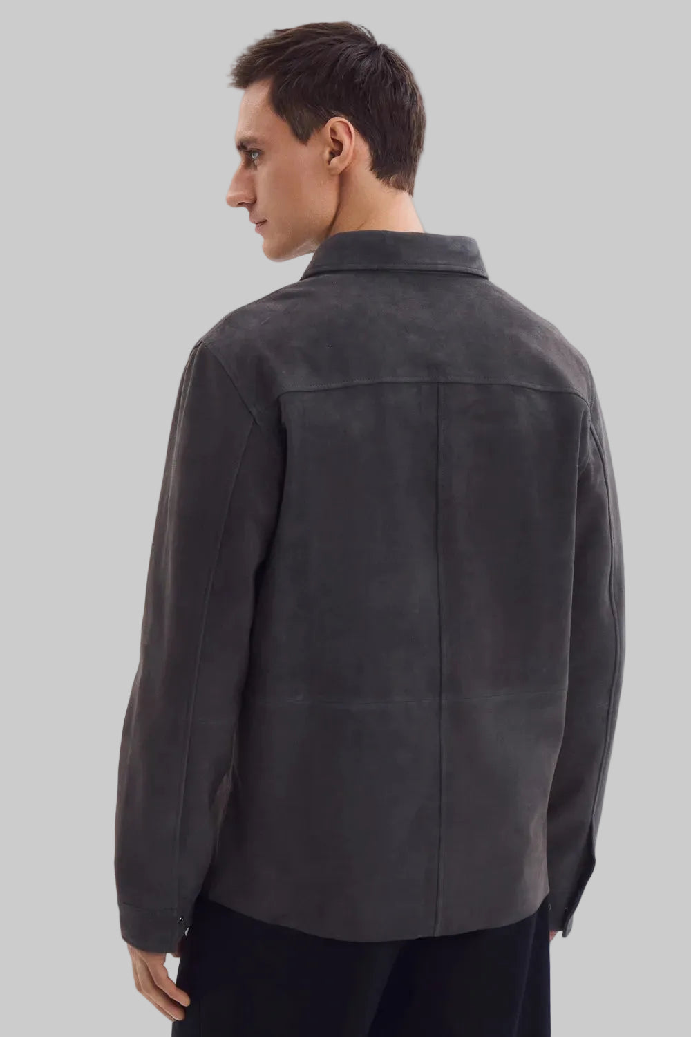 Front studio portrait of a man wearing a minimalist, charcoal gray suede shirt jacket snapped completely closed, featuring a clean concealed placket and point collar against a solid white background.